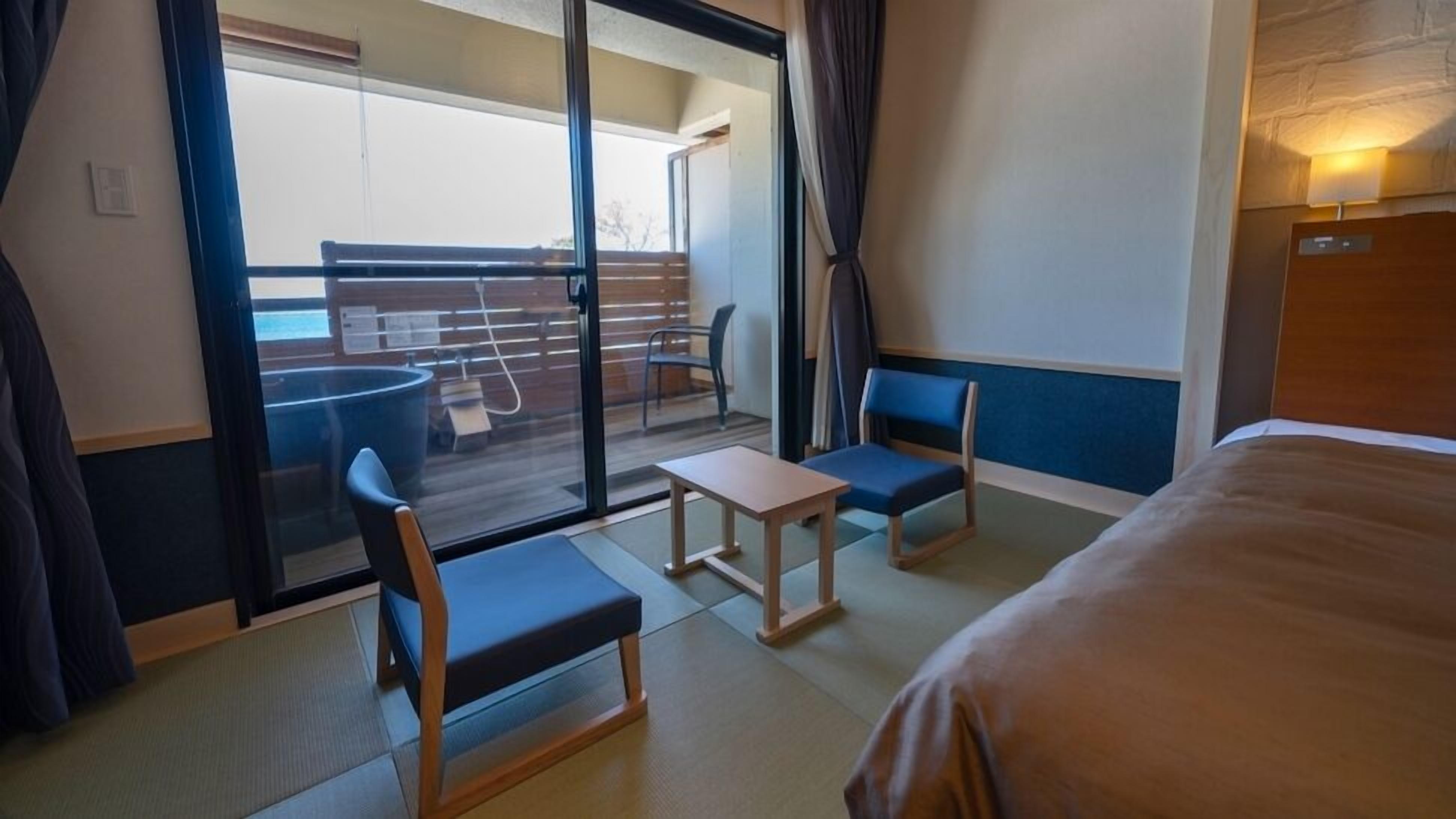 Une chambre d’hôtel comprenant un lit, deux chaises, une petite table et une porte-fenêtre coulissante donnant sur un balcon.