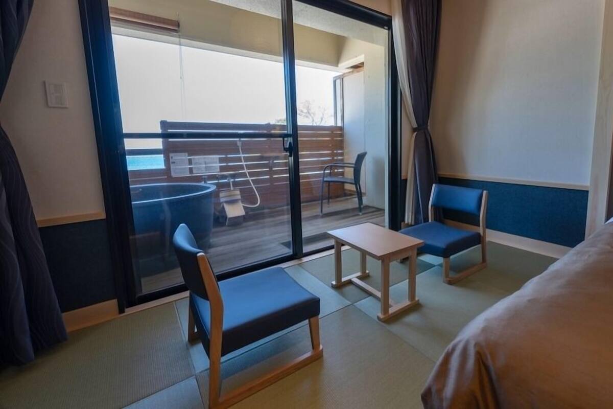 Quarto de hotel com cama, duas cadeiras, uma mesinha e porta de vidro que se abre para uma varanda.