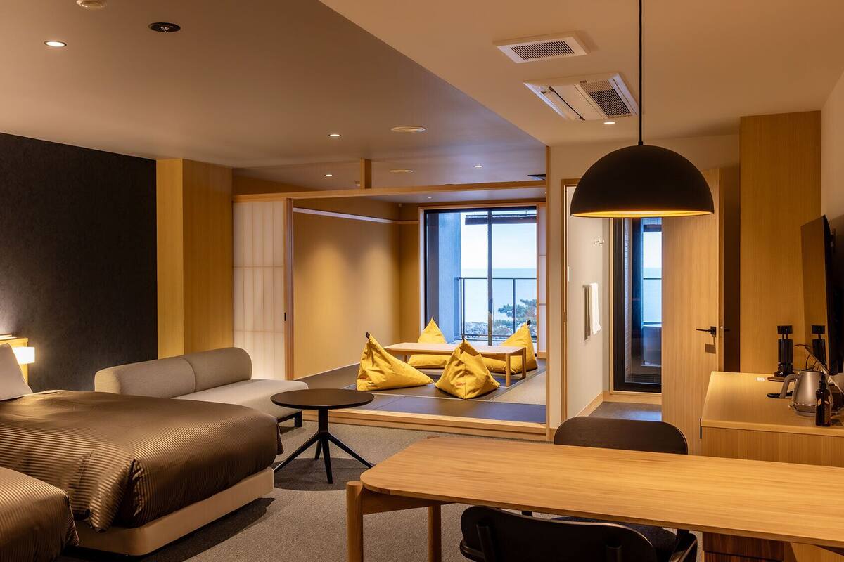 Quarto de hotel moderno com cama, sofá, mesa de jantar e vista para o exterior.
