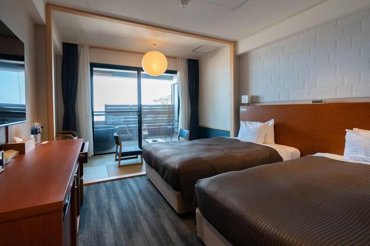 Quarto de hotel com duas camas, uma escrivaninha, uma cadeira, uma janela grande e vista para o exterior.