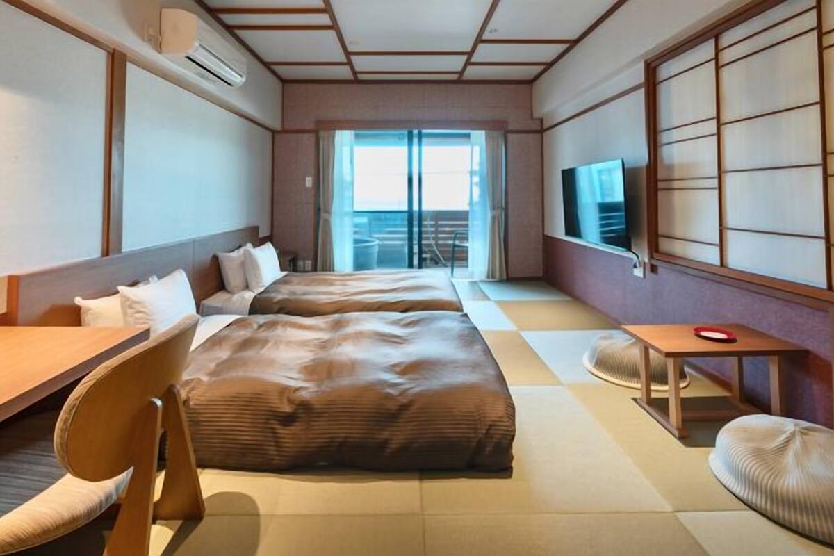 Quarto de hotel com cama, uma cadeira, uma mesinha, televisão e uma porta deslizante para uma varanda.