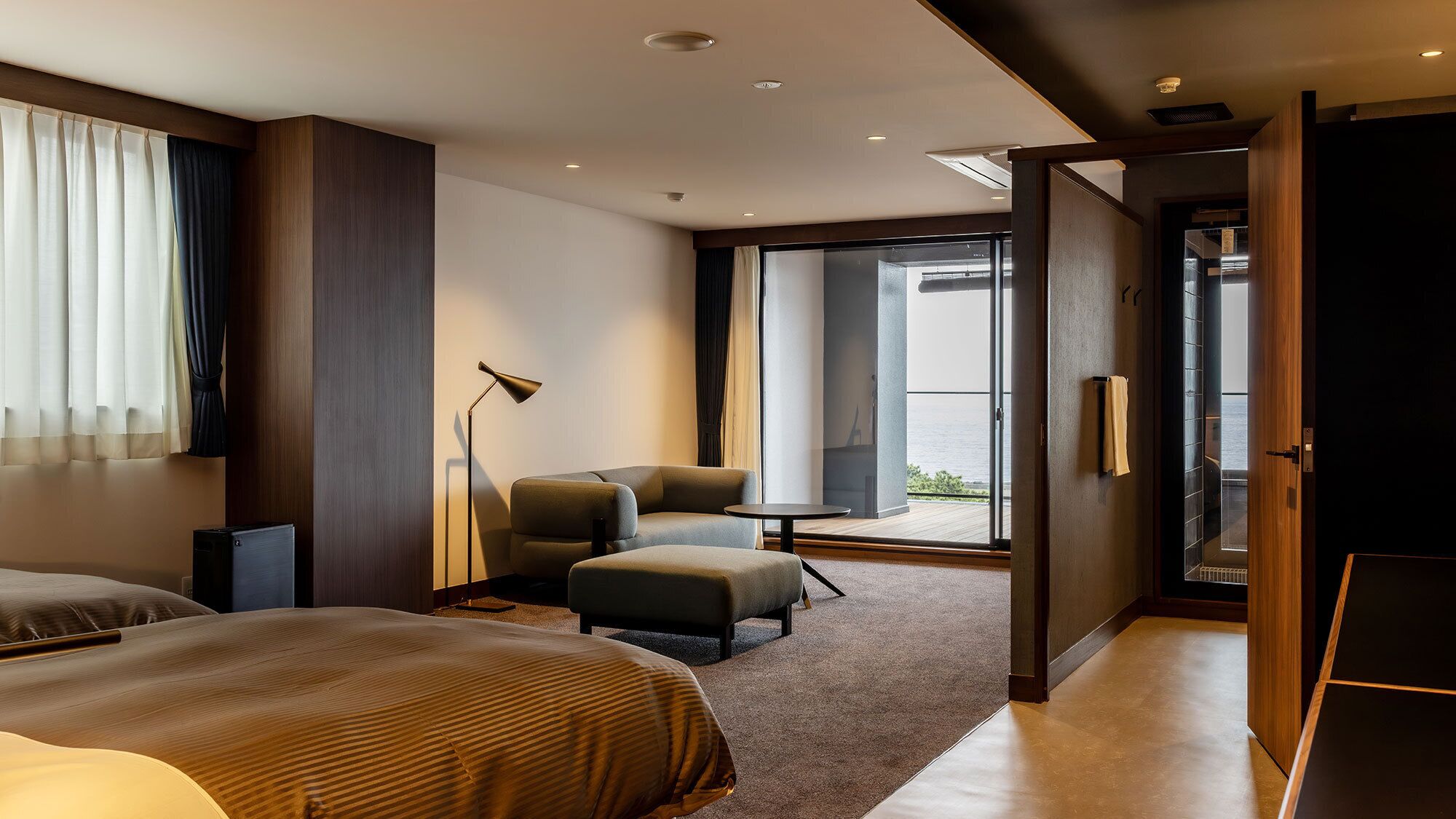 Une chambre d’hôtel moderne, équipée d’un lit, d’un coin salon avec une table, d’un lampadaire et offrant une vue sur l’extérieur grâce à une grande fenêtre.