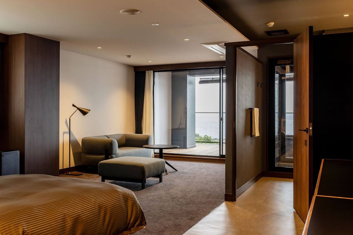 Quarto de hotel moderno com cama, área de estar com mesa, luminária de chão e vista pela grande janela.