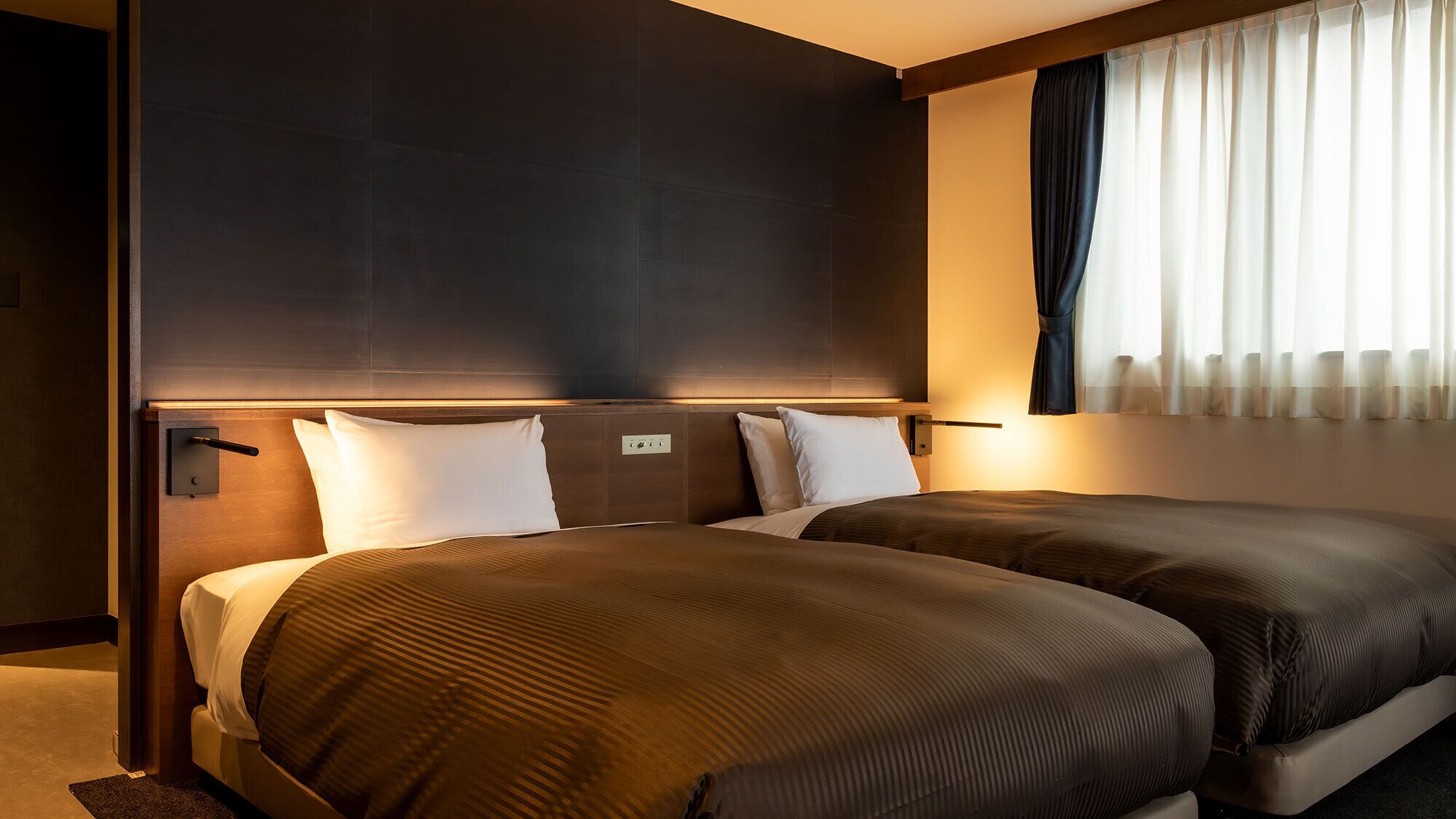 Une chambre d’hôtel avec deux lits, une tête de lit, une fenêtre avec des rideaux et un panneau de commande fixé au mur.