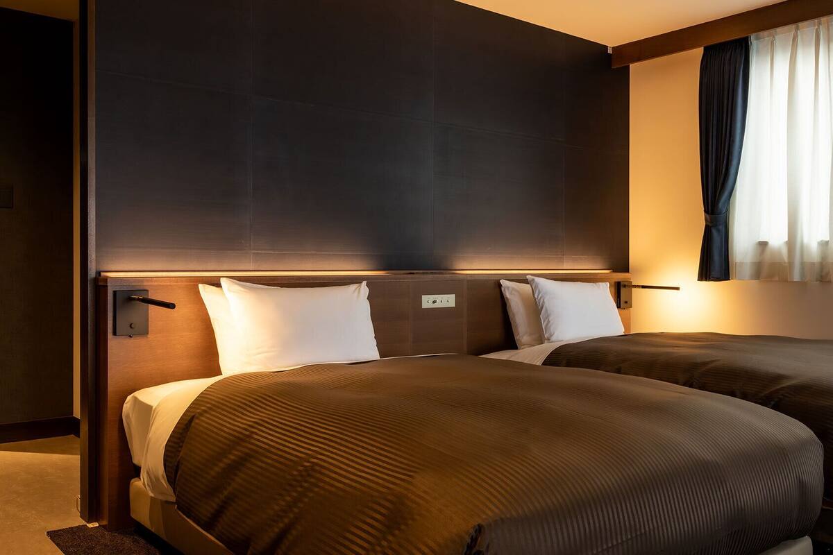Quarto de hotel com duas camas, cabeceira, janela com cortinas e painel de controle fixado na parede.