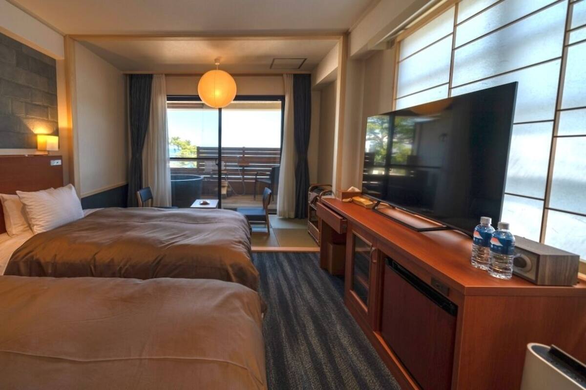 Quarto de hotel com duas camas, televisão, escrivaninha e varanda com área de estar ao ar livre.