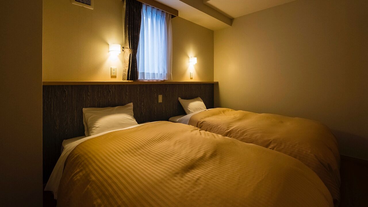 Une chambre d’hôtel avec deux lits, une tête de lit en bois, une fenêtre avec des rideaux et des lampes fixées au mur.