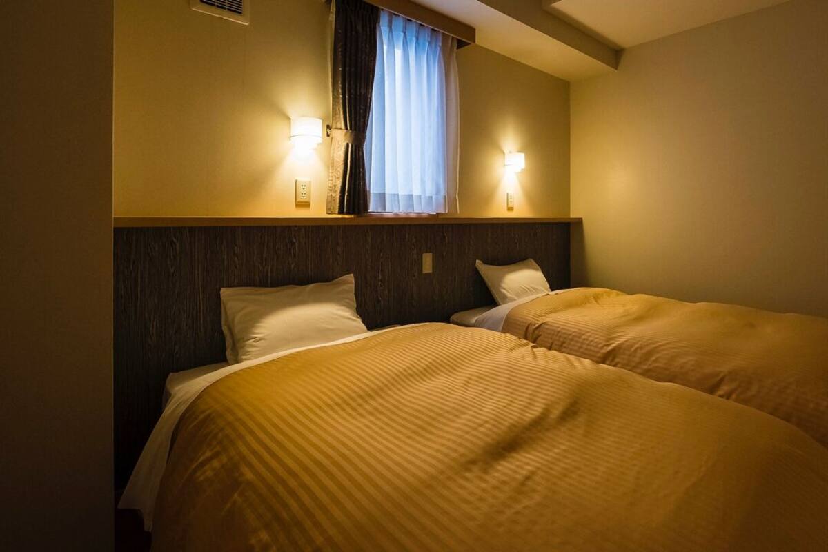 Quarto de hotel com duas camas, cabeceira de madeira, janela com cortinas e luminárias fixadas na parede.