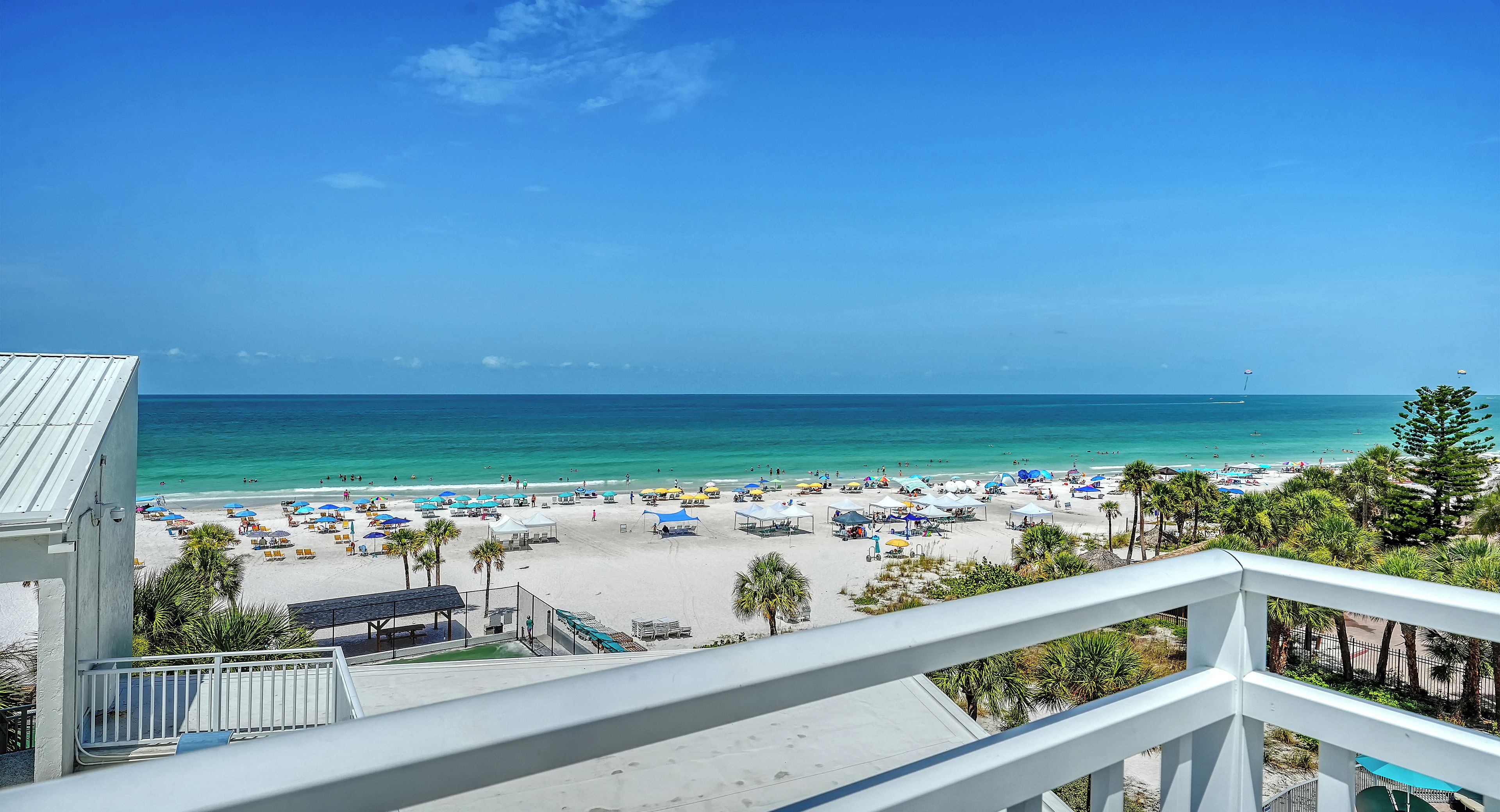 A beach view from a balcony with a railing, palm trees, and a clear sky.