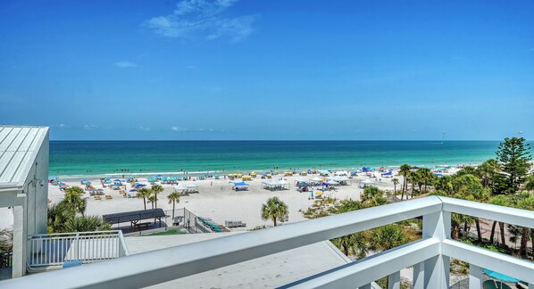 A beach view from a balcony with a railing, palm trees, and a clear sky.