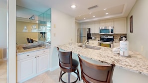 A kitchen with white cabinets, a stainless steel refrigerator, a microwave, an oven, and a sink. There are two wooden chairs and a marble countertop.