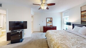 Quarto de hotel com cama, televisão, penteadeira, ventoinha no teto e uma janela grande.