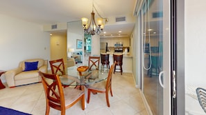 A dining area with a glass table and chairs, a chandelier, and a kitchen in the background.