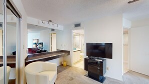 A modern bathroom with a shower, toilet, and sink. There is a white chair and a flat-screen TV mounted on the wall.