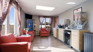 Une chambre d’hôtel compacte comprenant un coin cuisine, un espace salon avec un canapé et un fauteuil rouges, une petite table avec un vase, et une vue sur l’extérieur par une fenêtre agrémentée de rideaux.