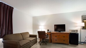 Une chambre d’hôtel avec un canapé marron, un bureau en bois avec une télévision, un petit réfrigérateur et une lampe.