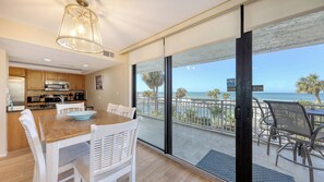A modern kitchen with stainless steel appliances, a dining area with a wooden table and white chairs, and a balcony with a view of the beach and palm trees. - House Of The Sun #216GF (Sarasota)