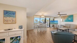 A spacious living area with a balcony overlooking a beach, a dining area, and a seating area with a television.