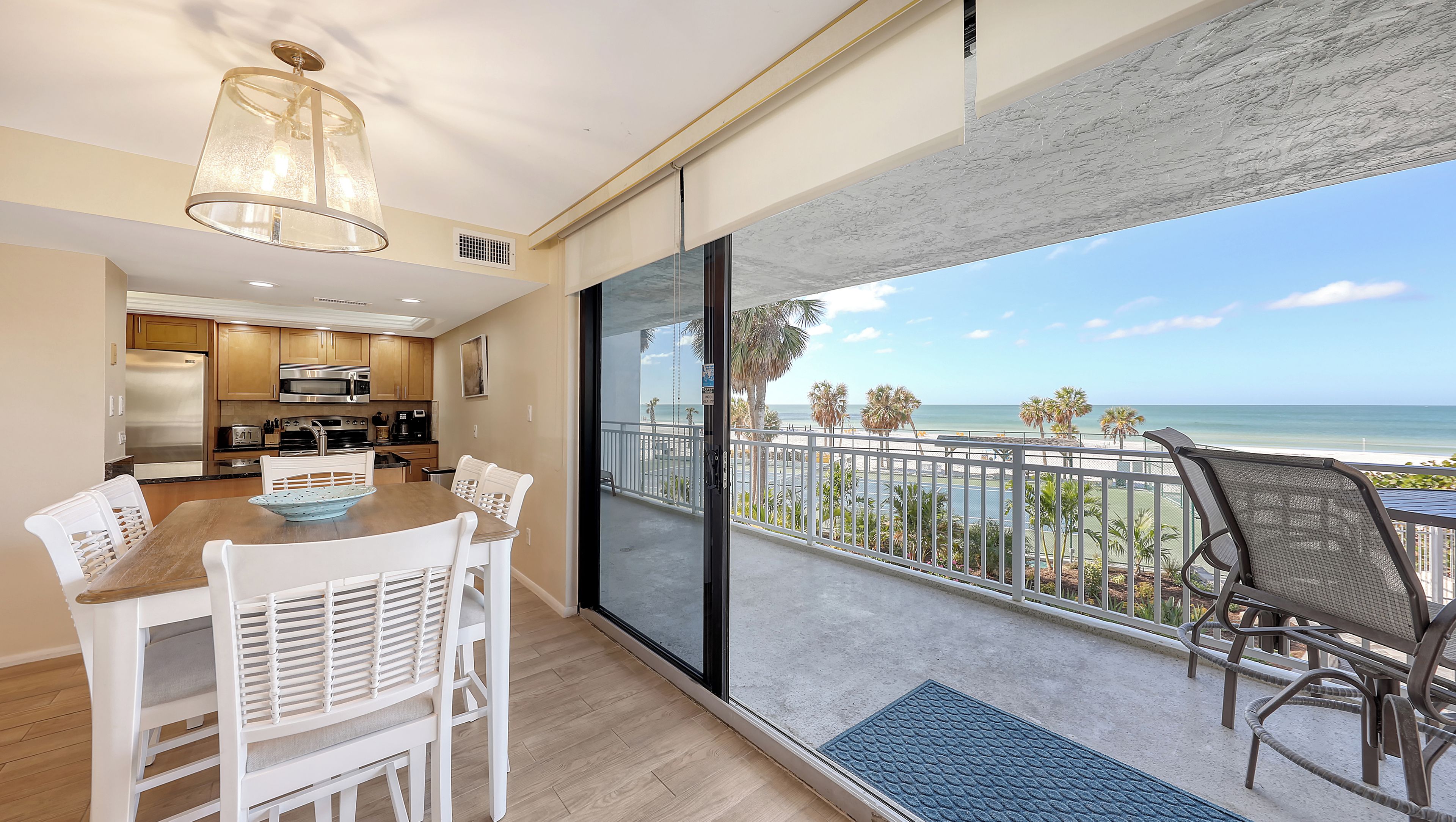 A modern kitchen with stainless steel appliances, a dining area with white chairs, and a balcony with a view of the beach and ocean.