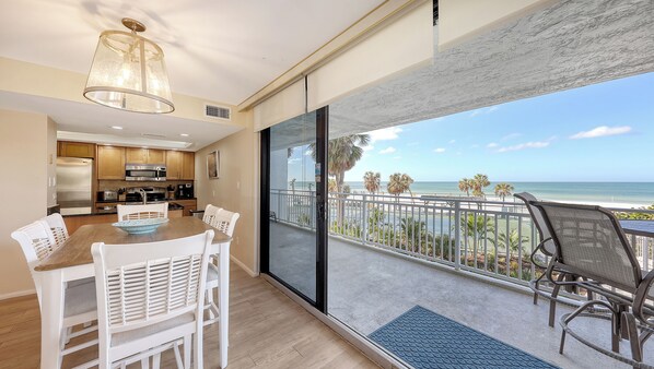 A modern kitchen with stainless steel appliances, a dining area with white chairs, and a balcony with a view of the beach and ocean.