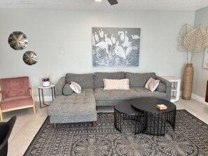 A living room with a grey sofa, a matching ottoman, a round coffee table, a red armchair, and decorative wall art. - Gulf View Condo with On-site Management - 411 (Sarasota)