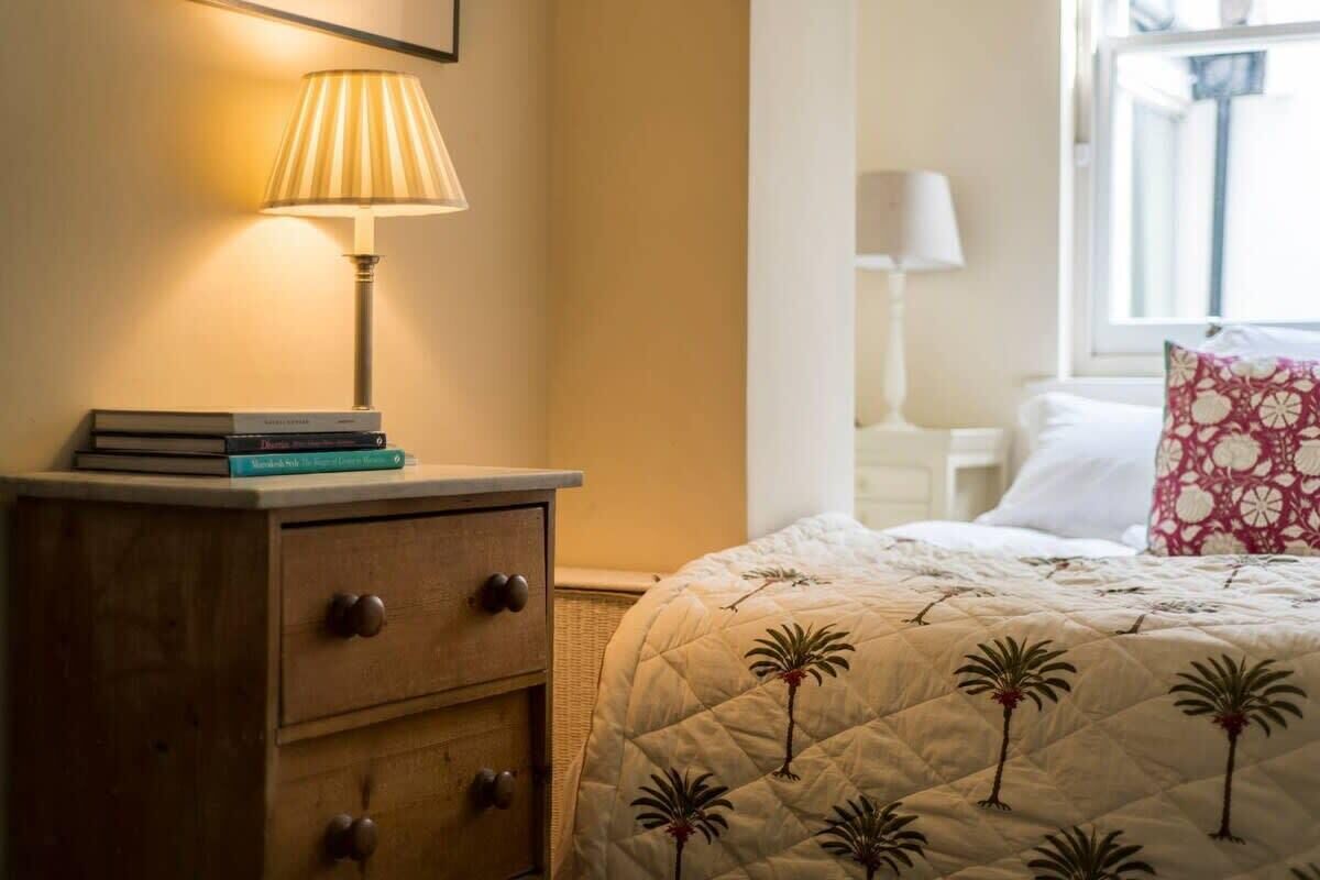 Een net opgemaakt bed met een gewatteerde spreidek die palmboommotieven toont, een bedlampje en een houten nachtkastje met een stapel boeken.