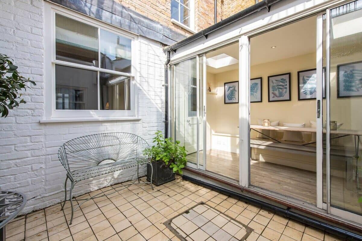 Un patio con una silla de metal, una planta en maceta y una puerta corrediza de vidrio que da a un espacio interior con una mesa y sillas.