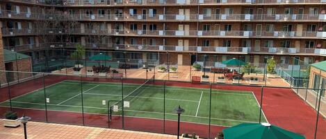 A multi-story hotel with a tennis court, outdoor seating, and a fenced patio area.