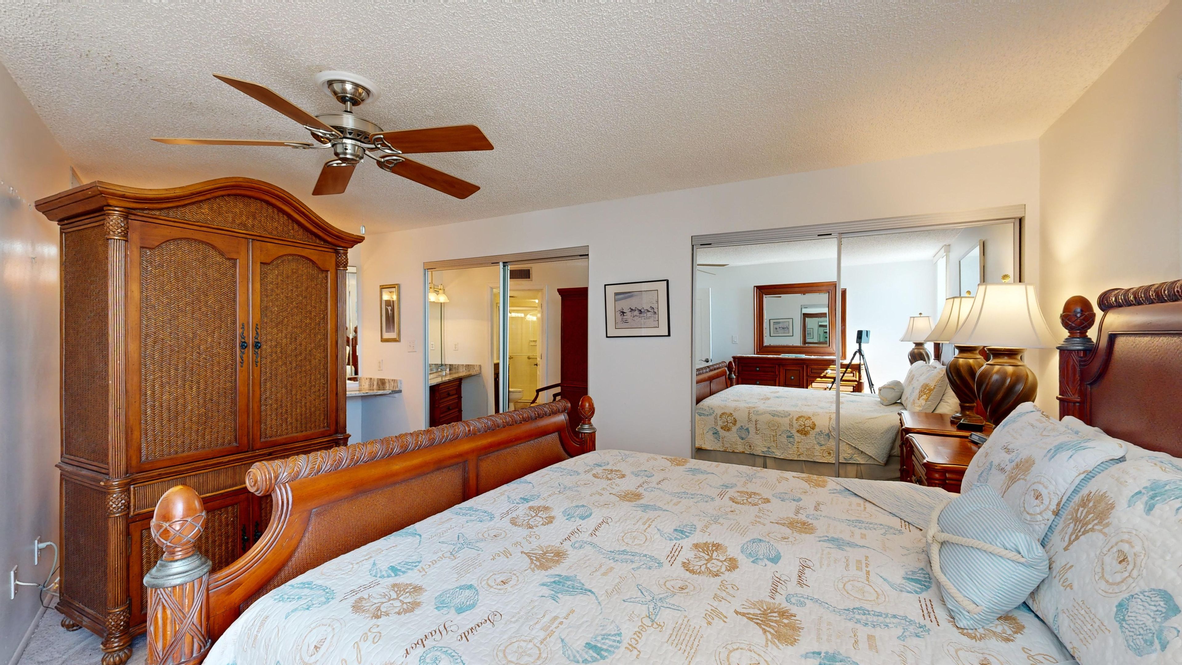Quarto de hotel com uma cama grande, um guarda-roupa de madeira, um ventilador de teto, um espelho e um banheiro visível através de uma porta aberta.