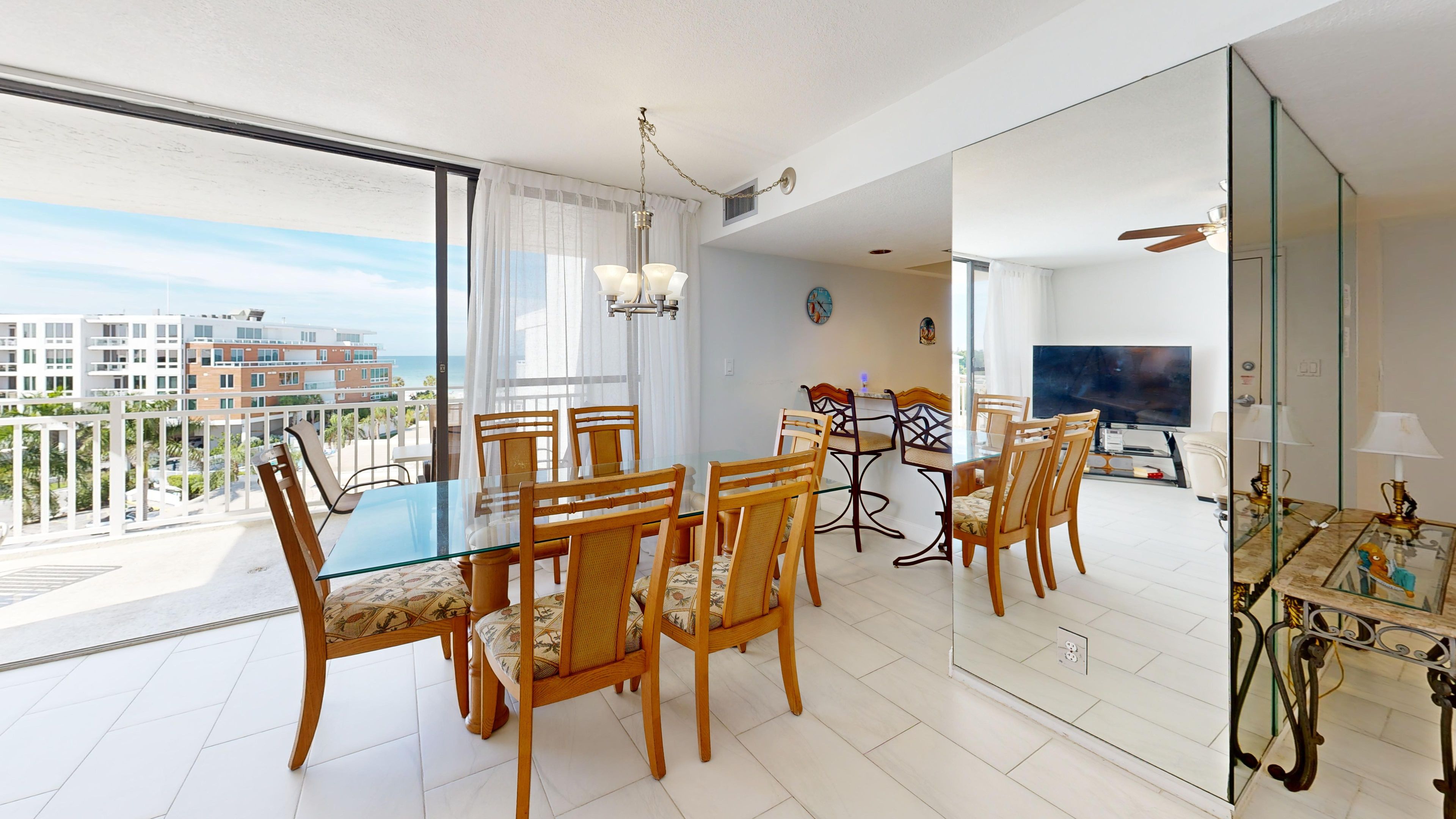 A dining area with a glass table and wooden chairs, a balcony with a view of buildings and the sea, and a living area with a TV and furniture.