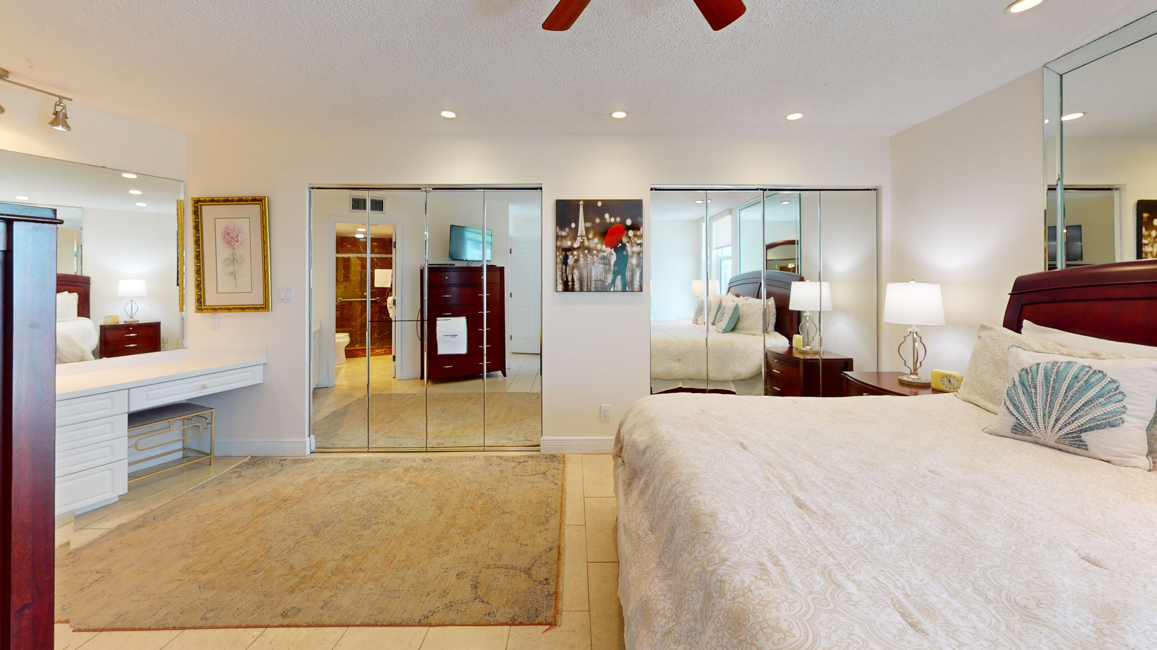 Quarto de hotel com cama, escrivaninha, espelho e banheiro visível por uma porta de vidro.