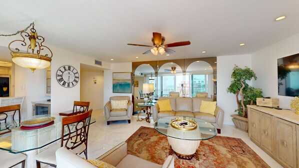 A spacious living room with a glass coffee table, a ceiling fan, and a large clock on the wall.