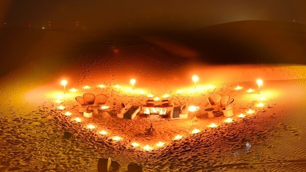 Disposição em formato de coração com velas e cadeiras sobre areia, sugerindo um ambiente romântico ou festivo.