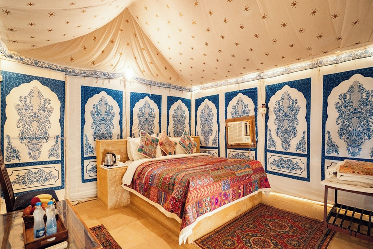 A room with a bed, a nightstand, a table with bottles, and a patterned rug.