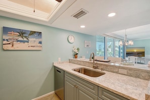 A kitchen with a sink, countertop, and cabinets. There is a clock on the wall and a painting of a beach scene. - House Of The Sun #316GF (Sarasota)
