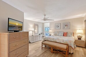 A bedroom with a television, a bed with decorative pillows, a bedside table with a lamp, and a wicker chest of drawers.