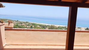 Un balcon abrité, doté d’un sol carrelé et de poutres en bois, qui offre une vue sur un paysage côtier avec des bâtiments et une plage.