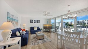 A modern living room with a large TV, two sofas, a dining table, and a balcony with a view of buildings. - House Of The Sun #303GS (Sarasota)