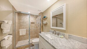 A modern bathroom with a glass-enclosed shower, a toilet, a sink with a marble countertop, and a large mirror. - House Of The Sun #303GS (Sarasota)