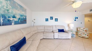 A living room with a beige sectional sofa, a blue accent pillow, a glass coffee table, and a ceiling fan. - House Of The Sun #312GV (Sarasota)