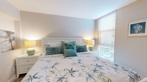 A bedroom with a bed, two bedside tables, a nightstand, a window with blinds, and wall art. - House Of The Sun #312GV (Sarasota)