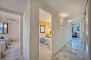 A hallway leading to a bedroom with a bed, a nightstand, and a lamp. There is a door on the left and a window with curtains in the bedroom. - RDR165 - Apt. Puerto Banús (Marbella)
