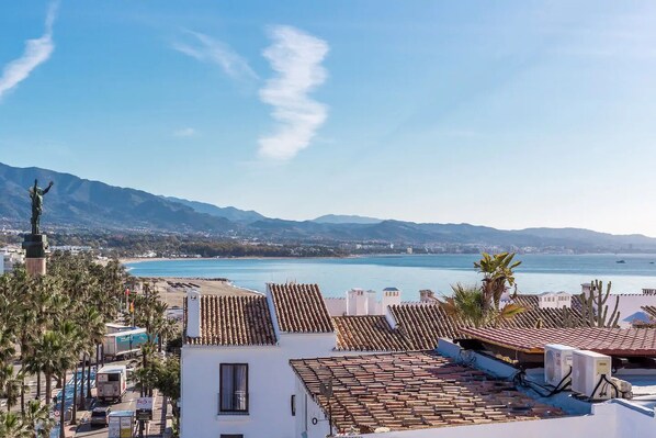 A coastal town with terracotta-roofed buildings, a statue, and a view of the sea and mountains. - RDR165 - Apt. Puerto Banús (Marbella)