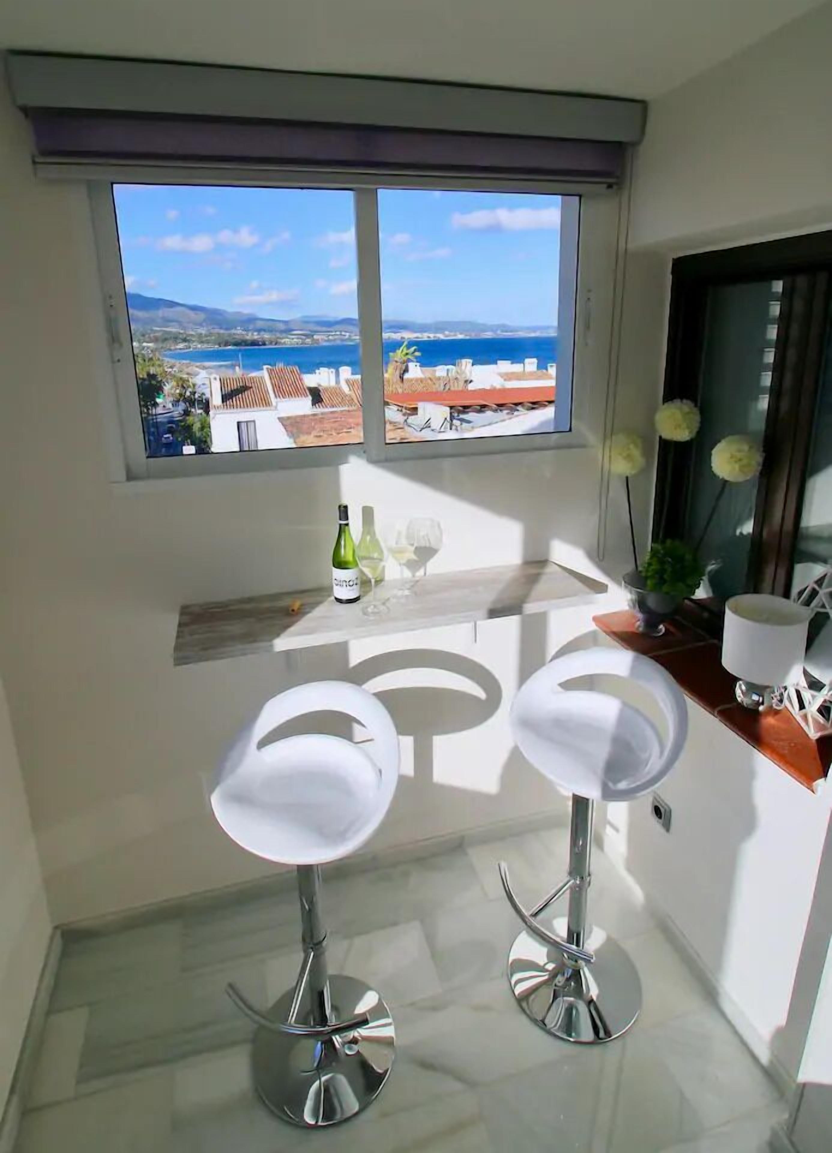 Una cocina moderna con dos taburetes de bar, un ventanal con vista al mar y una pequeña encimera con una botella y vasos.