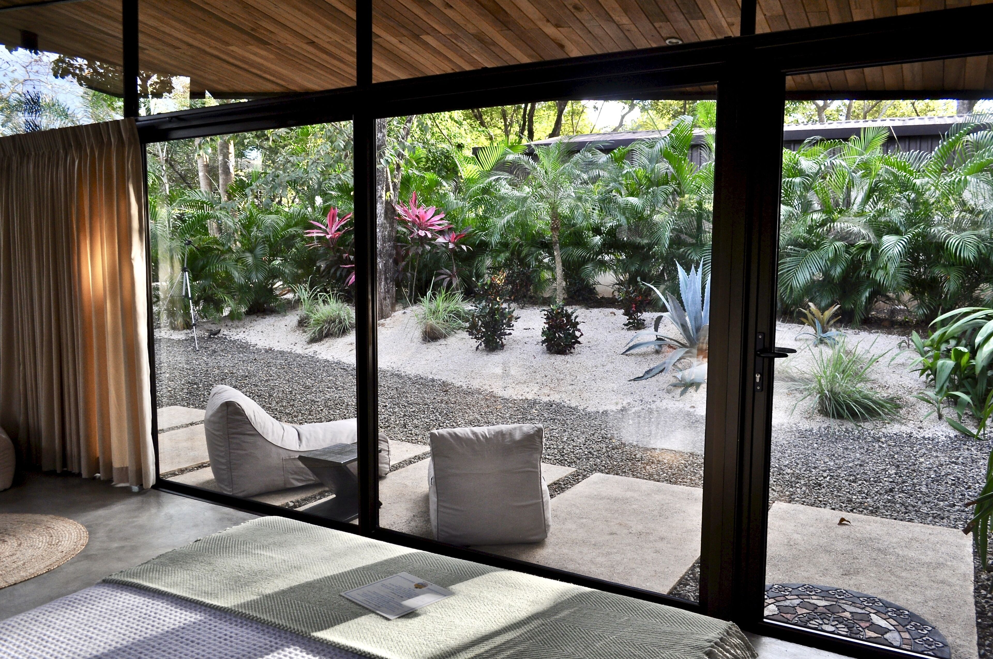 Una habitación con un amplio ventanal corredera que da a un jardín con diversas plantas y una zona de estar con cojines.