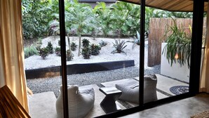 A view from a room with large windows overlooking a garden with various plants and a paved area with seating. - Villas Kalei (Cabo Velas)