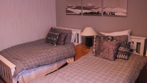 Un dormitorio con dos camas, cada una con ropa de cama y almohadas estampadas. Hay una mesita de noche con una lámpara entre las camas. Las paredes están pintadas de un color claro, y hay tres cuadros de paisajes sobre las camas.