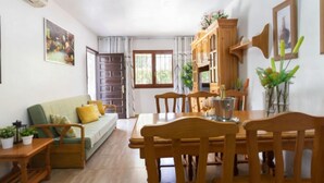 Un salon chaleureux avec un canapé, une table à manger en bois et des chaises. On y trouve une fenêtre avec des rideaux, une porte et une petite table ornée de plantes.