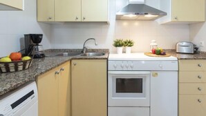 Une cuisine compacte équipée d’une cuisinière, d’un four et d’un évier. On y trouve des placards, une machine à café et un panier de fruits sur le plan de travail.