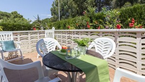 Une terrasse extérieure avec des chaises blanches et une table recouverte d’une nappe verte, le tout entouré d’une clôture blanche et d’une végétation luxuriante.